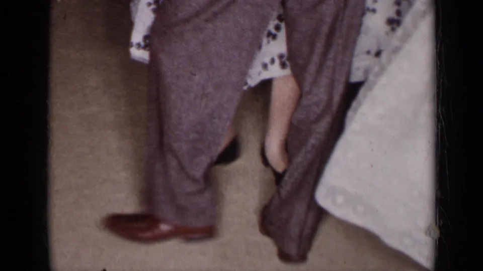 1954 New York home-movie frame focused on the couple's stepping feet, with brown dress shoes, gray trousers, and a light skirt edge.