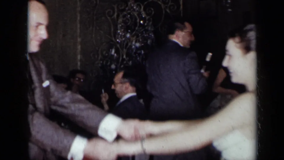 A suited man and woman dance close to the camera at the 1954 New York party, with seated guests and table settings behind them.
