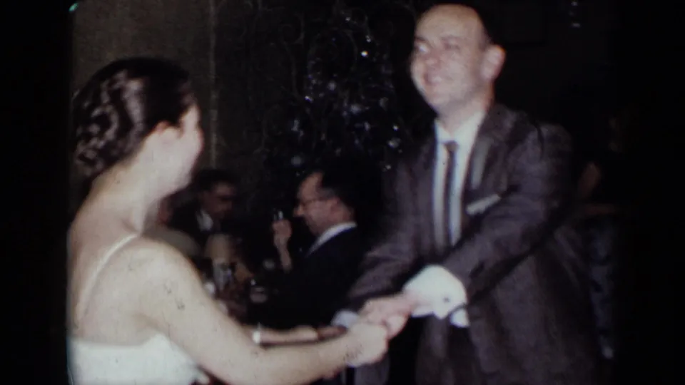 A woman with braided hair dances with a suited man in a dim 1954 New York party room, with diners and glassware visible behind them.