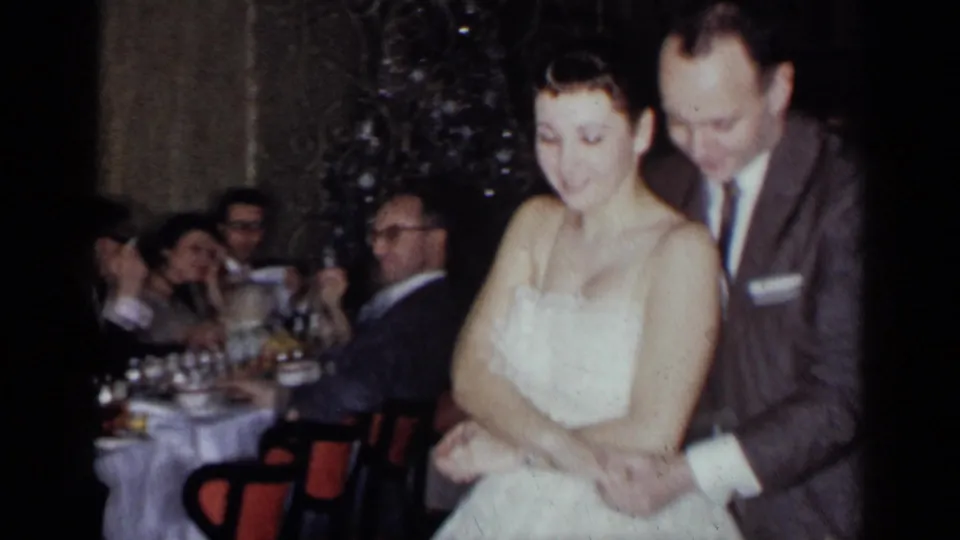 A suited man dances closely behind a woman in a white dress during the 1954 New York holiday party, with dinner guests in the background.