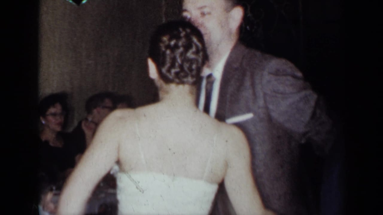 1954 New York reception frame showing a woman in a white strapless gown dancing with a suited man as guests sit behind them.