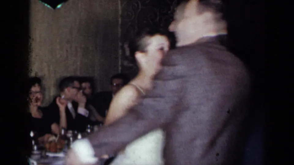 1954 New York home-movie frame of the couple turning on the reception floor with guests seated at tables behind them.