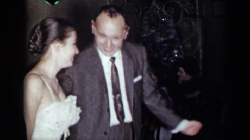 1954 New York frame showing a smiling woman in a white gown dancing beside a man in a suit and tie.