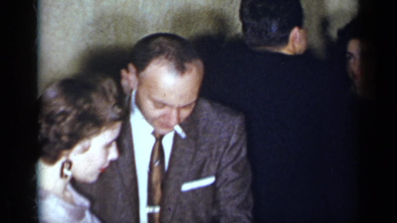 Man in a brown suit looks down beside a woman in a light purple dress at an indoor party.