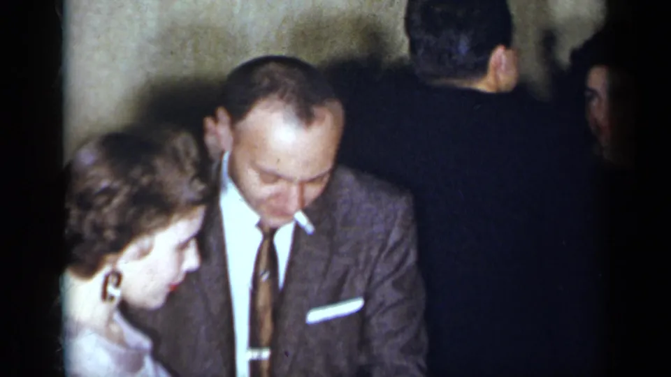 At a 1954 New York social gathering, a suited man and woman in a light purple dress stand close while other guests face away.