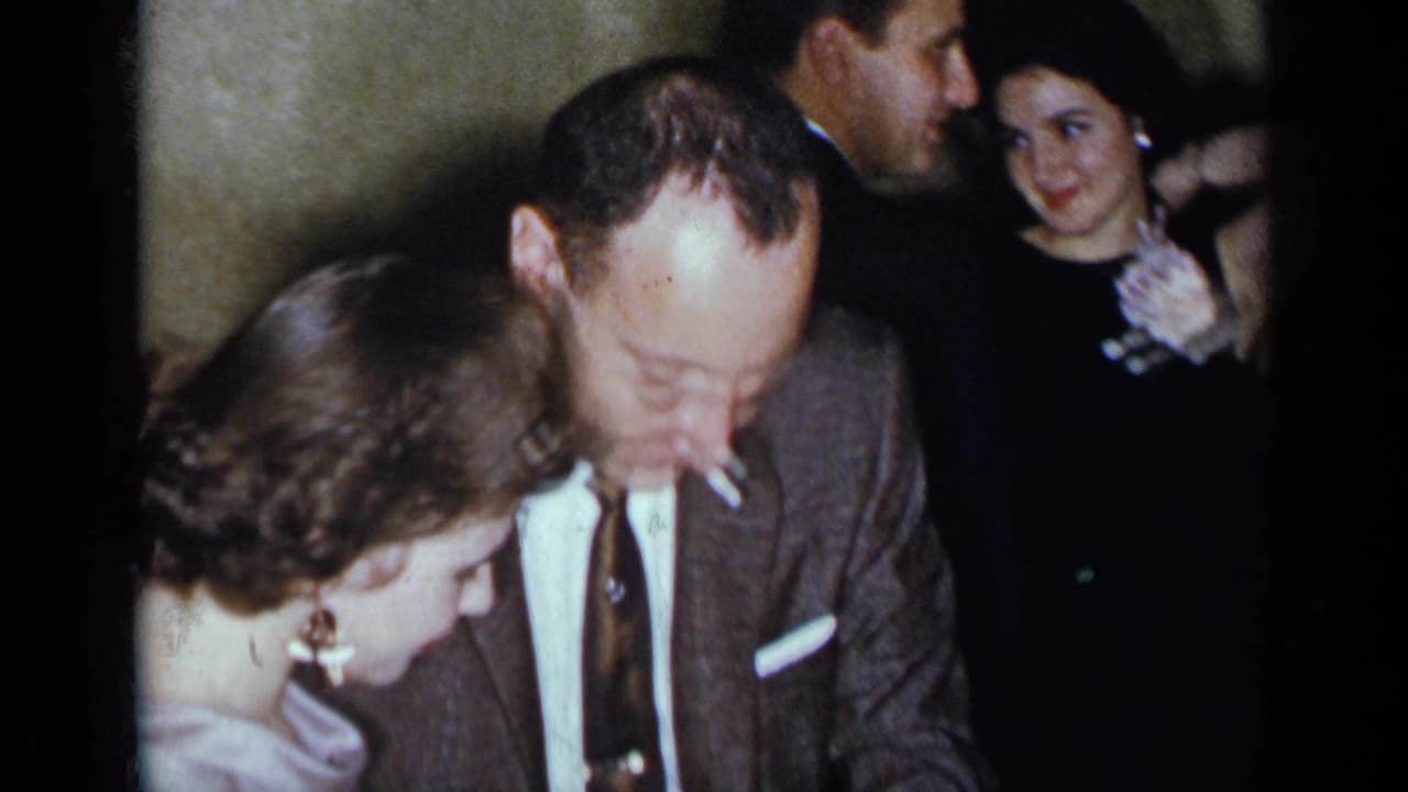 Man with a cigarette stands close to a woman in a light purple dress as guests gather behind them.