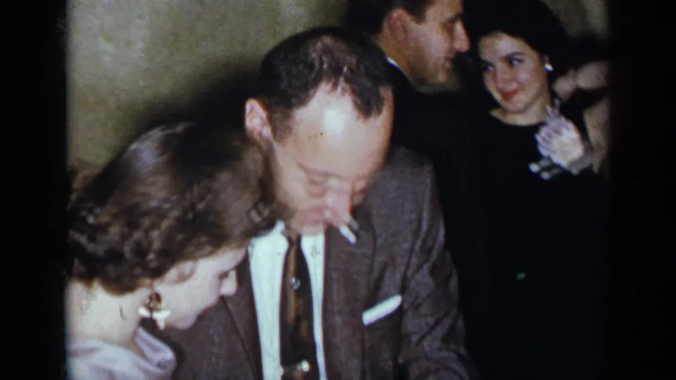 In the 1954 New York home-movie frame, a man in a suit holds a cigarette while standing close to a woman in a light purple dress.