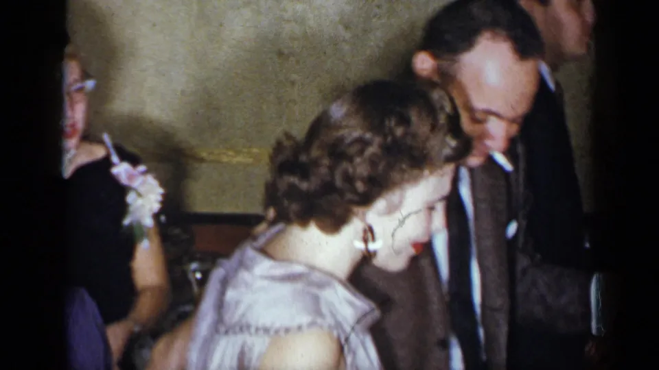 A 1954 New York party frame shows the woman in a ruffled light purple dress near the suited man with a cigarette, with guests around them.