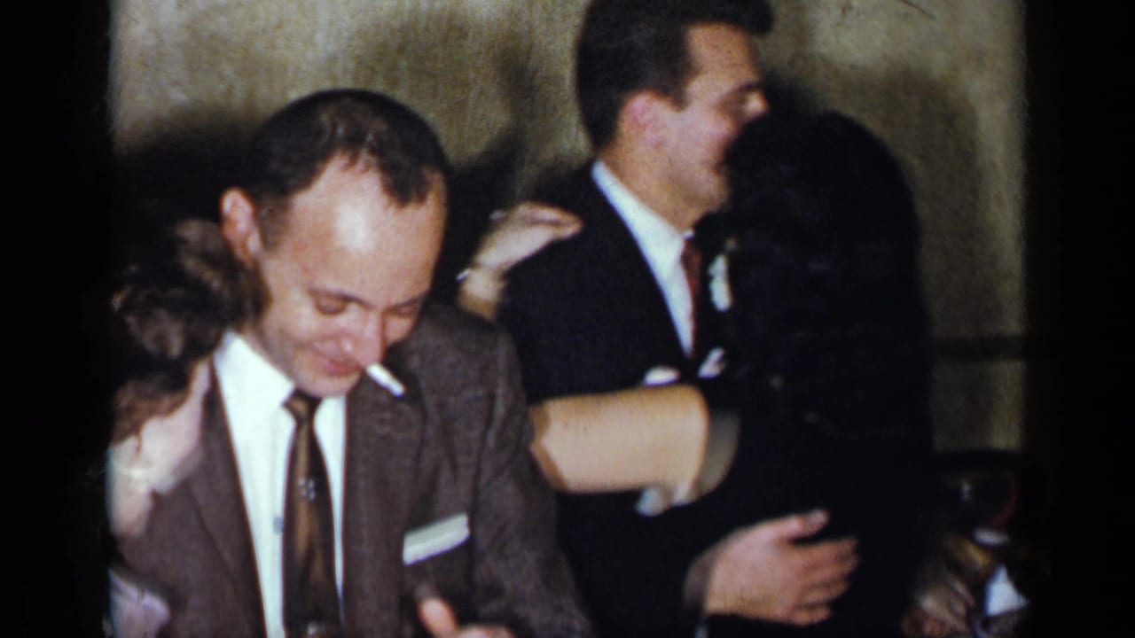 1954 New York home-movie frame of adults dancing closely indoors, including a man in a brown suit and partially visible partners.