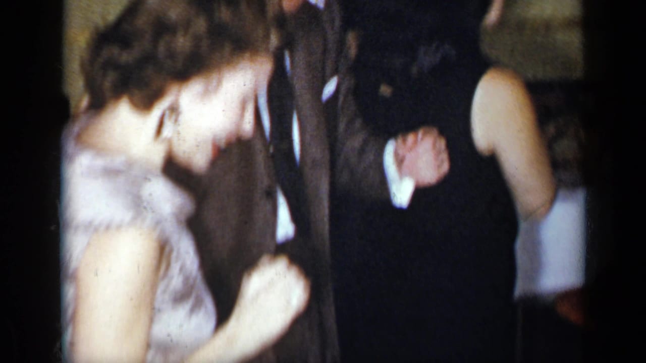 1954 New York home-movie frame showing a woman in a pale dress dancing with a suited man in a dim private interior.