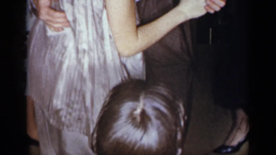 1954 New York home-movie frame of a couple dancing hand in hand in a dim interior, with nearby guests partly visible.