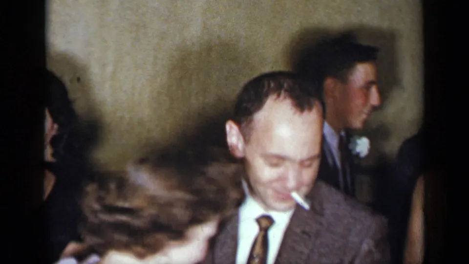 1954 New York home-movie frame showing a suited man smoking beside other formally dressed guests in a crowded interior.