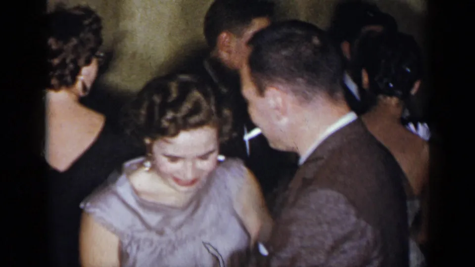 1954 New York home-movie frame of a smiling woman in a light dress dancing with a man in a brown suit among guests.