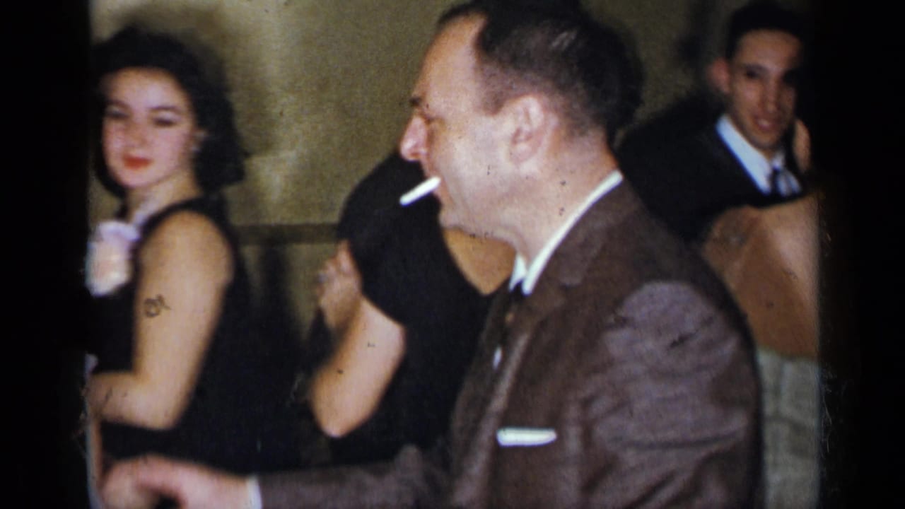 1954 New York home-movie frame of adults mingling indoors, with a woman in black, a smoking man in a brown suit, and guests in formal attire.