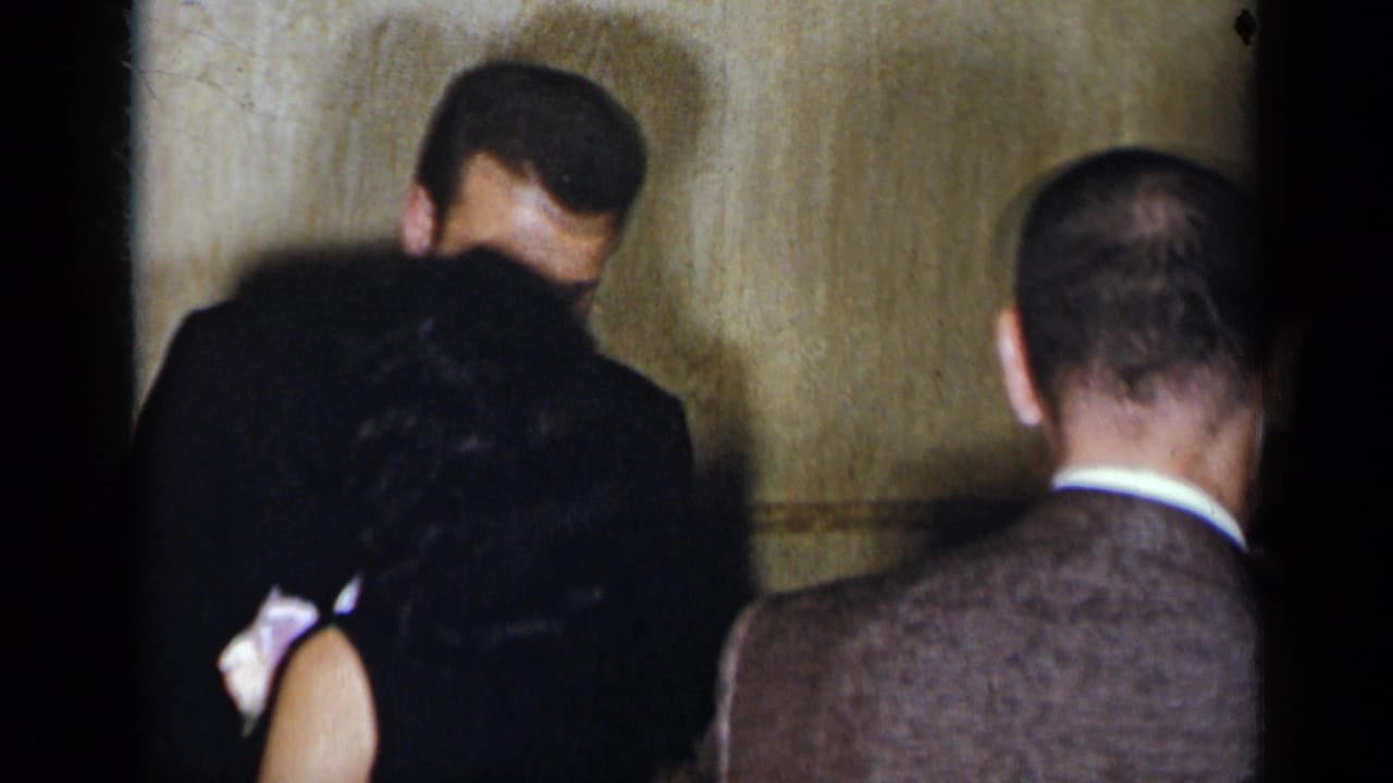 1954 New York party frame showing adults in suits and dark dresses gathered closely in a dim private interior.