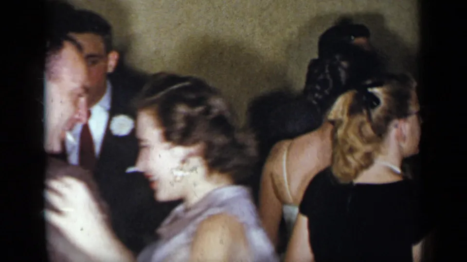 1954 New York home-movie frame with a woman in a light blue dress dancing among formally dressed adults in a crowded room.