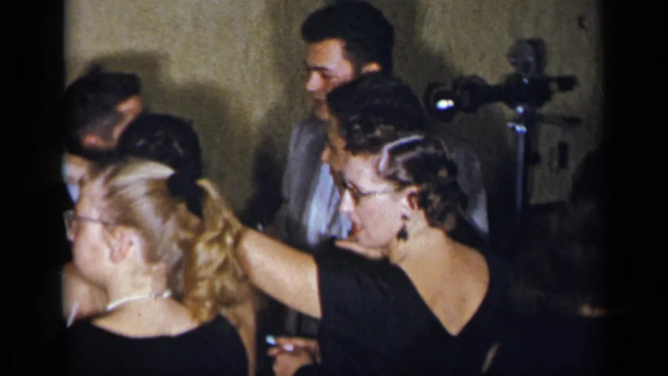 1954 New York indoor party frame showing adults in formal clothing, a woman with glasses, and a tripod-mounted camera near the wall.