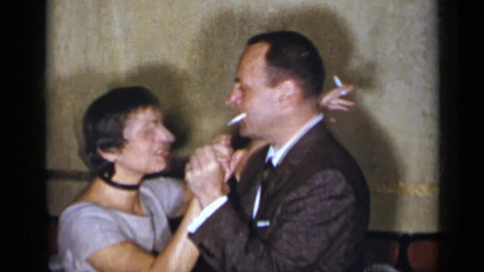 A man in a dark suit holds a cigarette while dancing closely with a woman in a light dress inside the 1954 New York gathering.