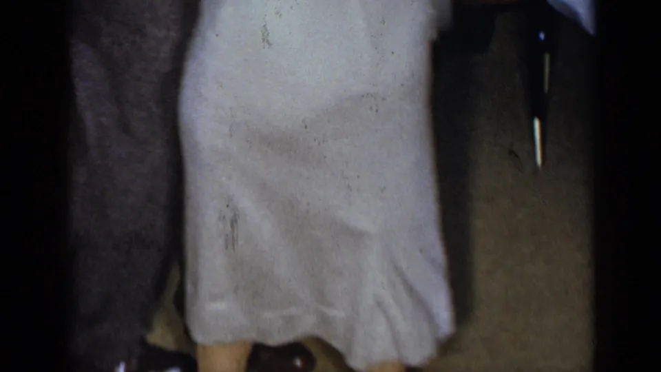 A cropped home-movie frame shows the dancers' lower torsos, a light dress, dark suit fabric, wall shadows, and part of the party room.