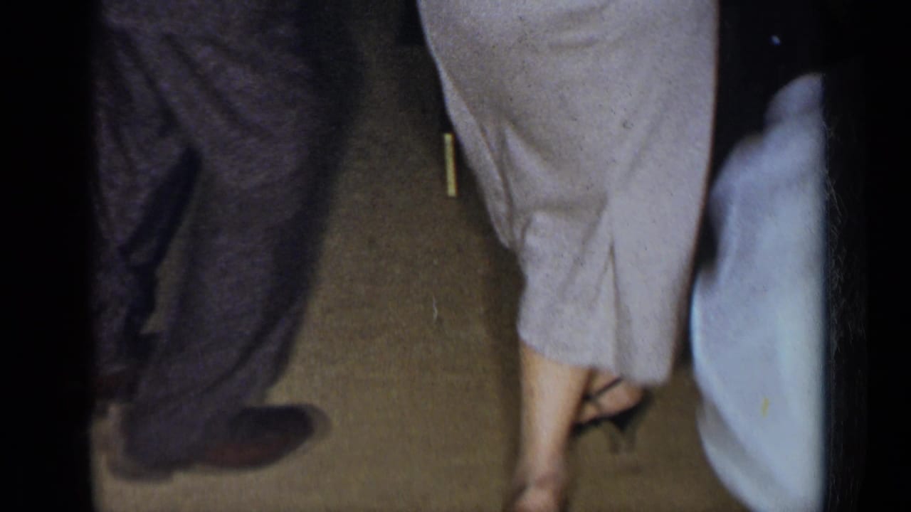 Lower bodies of adults dancing on a carpeted floor near chairs.