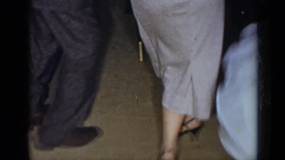 In a 1954 New York home-movie frame, adults in dress clothing move across a carpeted indoor party floor.