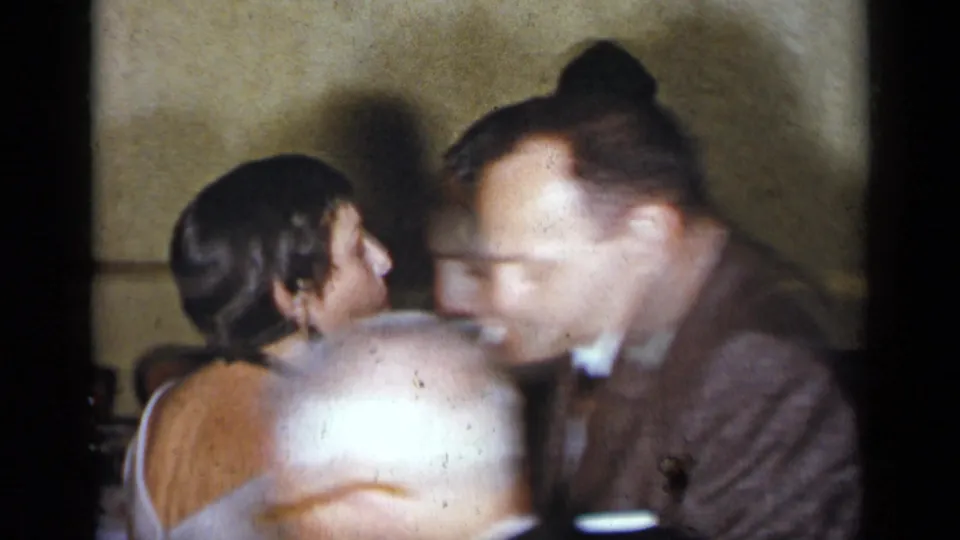 Adults dance and talk in a close, soft-focused 1954 New York party frame, partly blocked by a foreground guest.