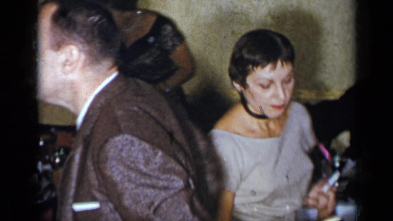 Adults at an indoor party, including a man in a brown suit and a seated woman holding an object.