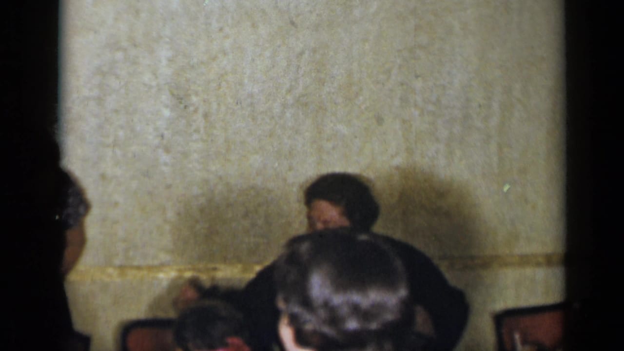 Adults partly obscured in a dim room with chairs and textured walls visible.