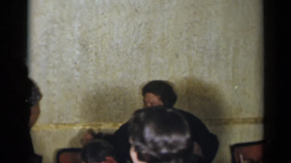 Dim 1954 New York home-movie frame with party guests partly obscured near chairs and textured interior walls.