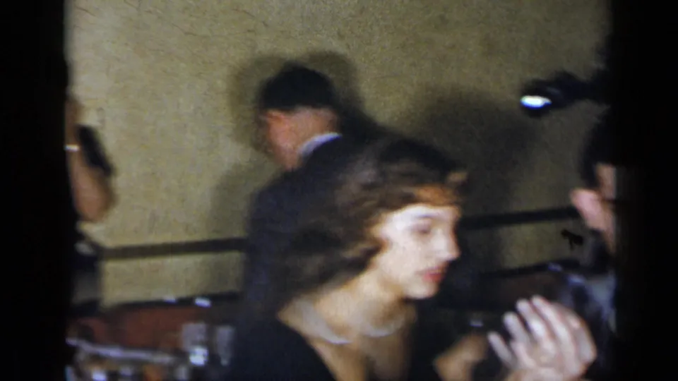1954 New York home-movie frame showing a woman seated in the foreground as other adults mingle in a private interior.