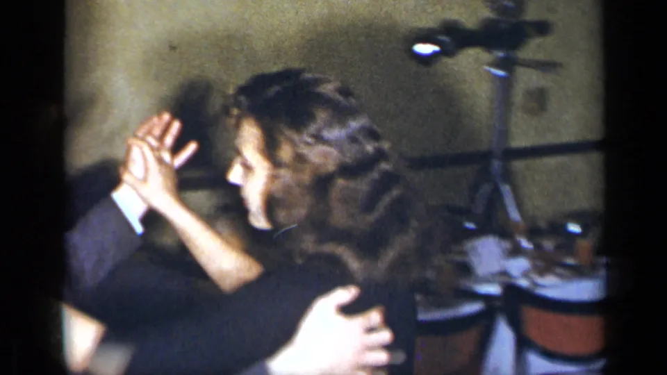 1954 New York home-movie frame of two adults dancing closely, with chairs and textured walls behind them.