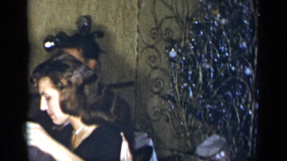 A woman in formal dress is seen in profile as the handheld 8mm camera passes other guests at the 1954 New York gathering.