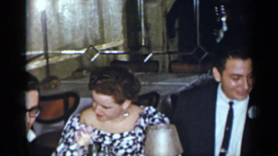 At the 1954 New York indoor event, a seated woman in a patterned dress appears beside a man in a dark suit and nearby table lamps.