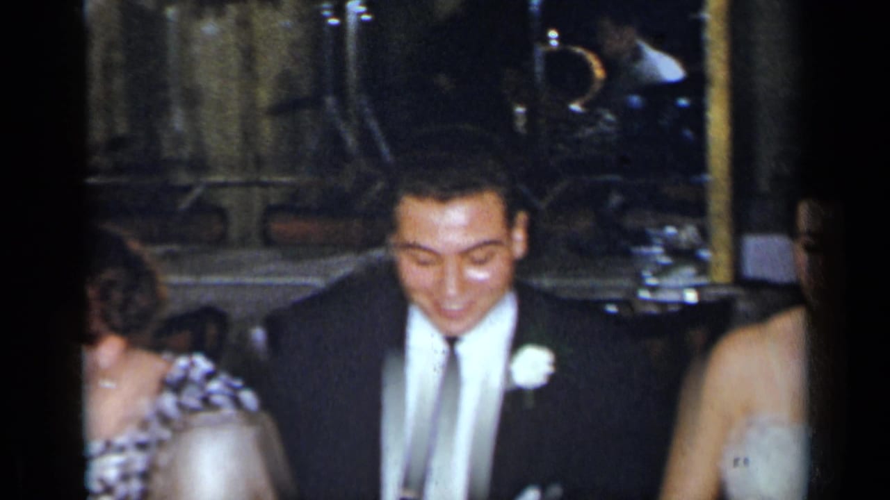 Groom in a dark suit sits beside guests at a dim restaurant table.