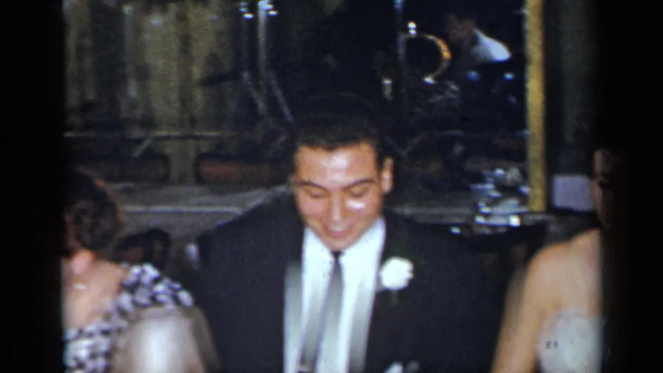 1954 New York home-movie frame of a groom in a dark suit with a white boutonniere seated beside guests in a dim reception interior.