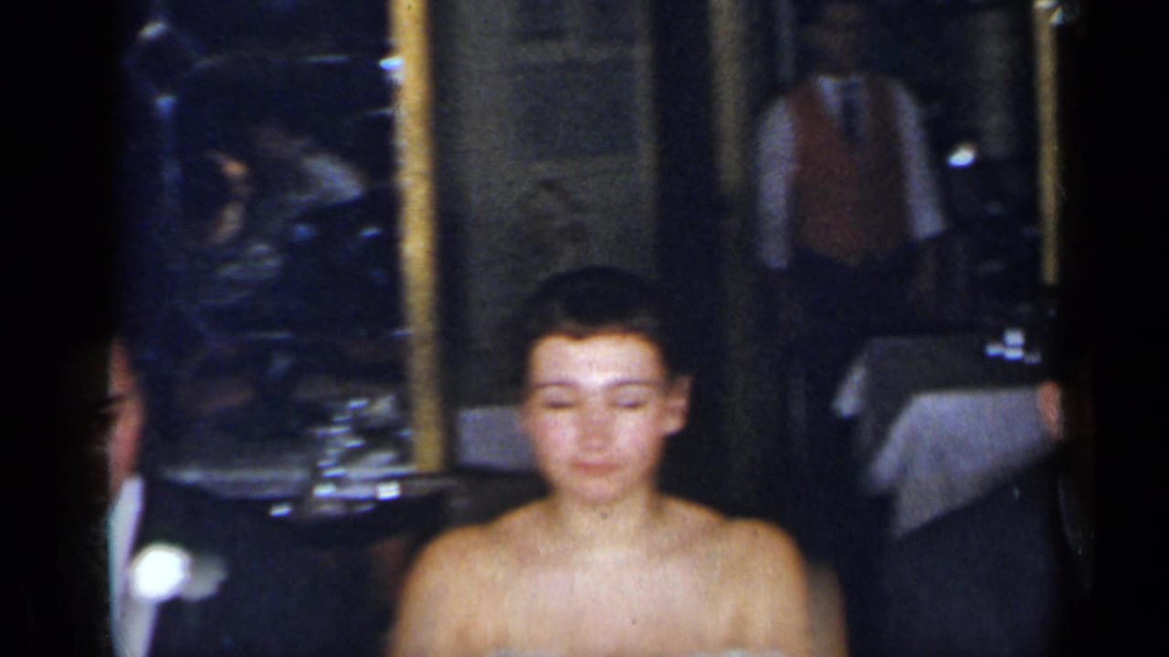 1954 New York reception frame showing a woman in a strapless formal dress near a suited guest and table settings in a dark restaurant interior.