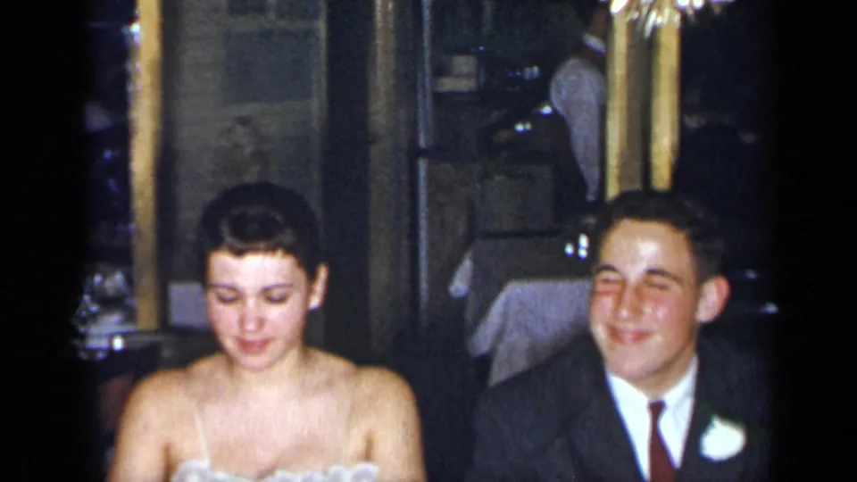 1954 New York home-movie frame of the bride in a white strapless gown and groom in a dark suit seated together at the reception.
