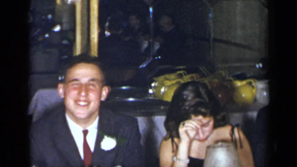 1954 New York reception frame with the groom smiling broadly beside a seated woman who covers her face in a candid moment.