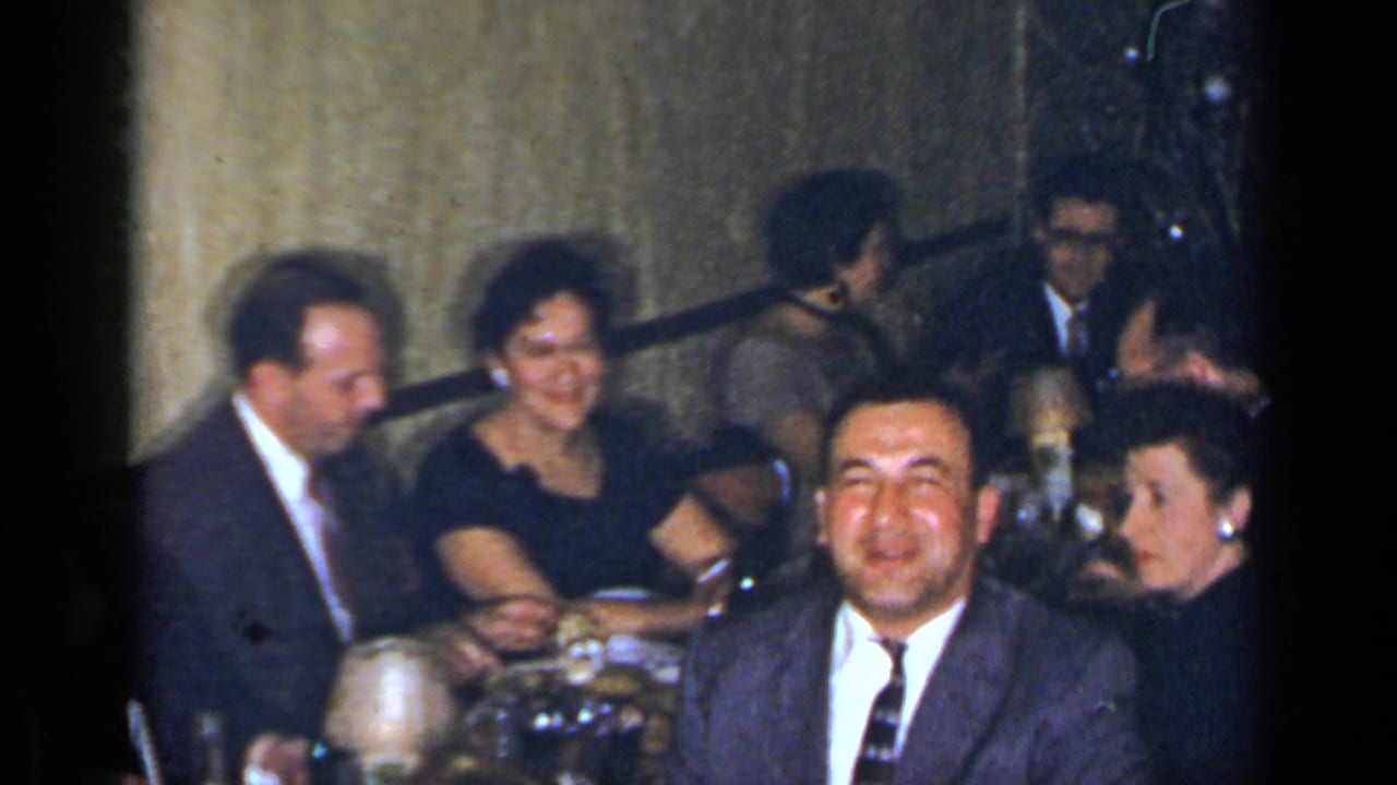 1954 New York home-movie frame showing formally dressed adults seated at a dining table during a meal.