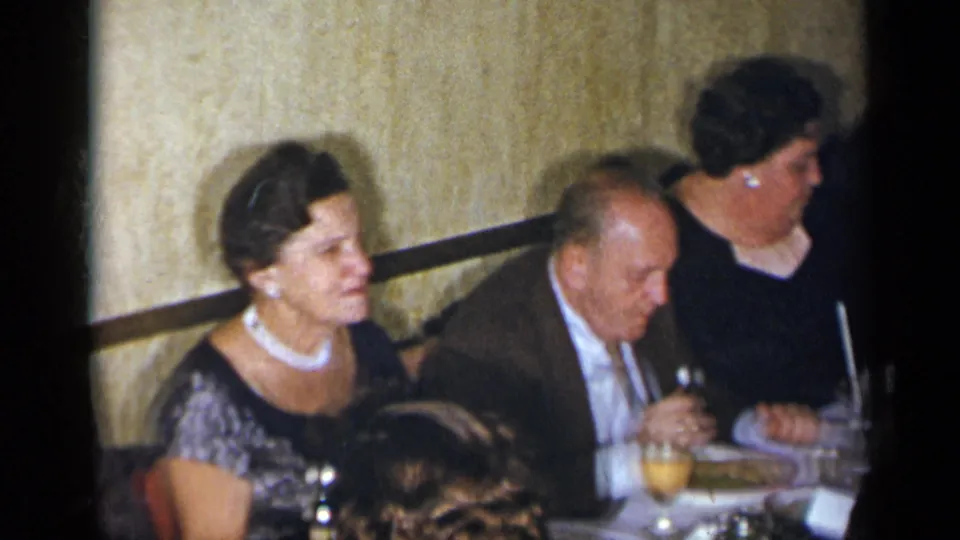 1954 New York home-movie still of adults at a formal dinner, including a woman in a dark dress and an older man eating.