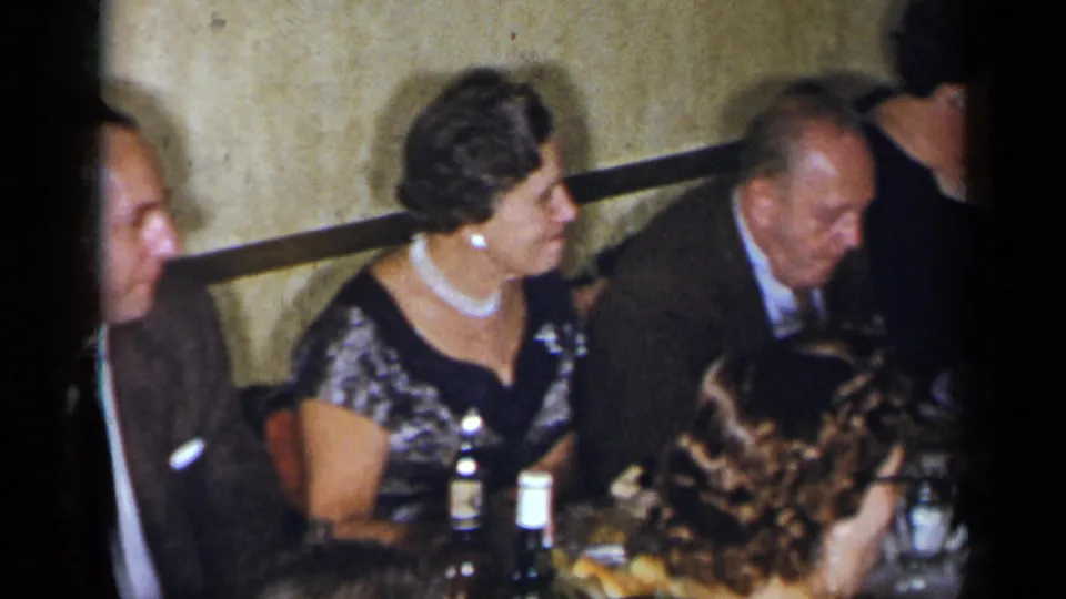 1954 New York dinner party frame showing adults in formal dress seated at a crowded table with bottles and glassware.