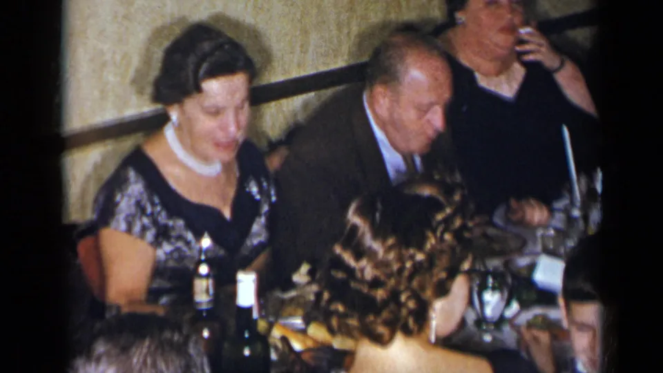 1954 New York home-movie frame showing a woman in a black and silver dress eating beside a suited man at a crowded dinner table.