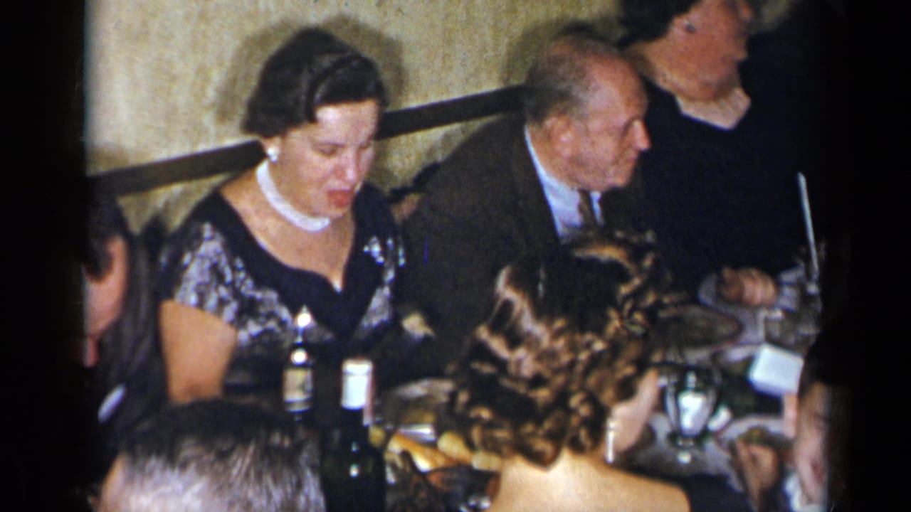 1954 New York frame of adults at a formal dinner, with a woman in a pearl necklace and a suited man visible among table settings.
