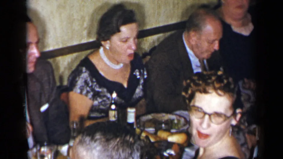1954 New York home-movie frame with a formally dressed woman, nearby suited men, and a guest with glasses at a dinner table.