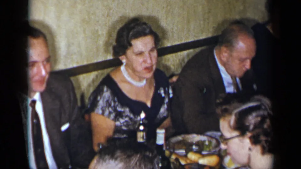 1954 New York frame showing a woman in a black and silver dress seated beside men in suits during a formal dinner gathering.