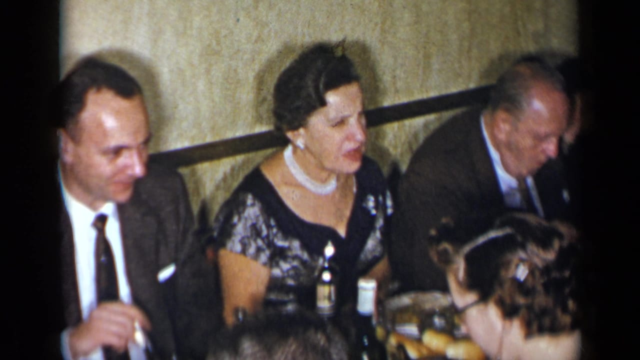 Adults seated at a dinner table with drinks, food, suits, and evening dress visible.