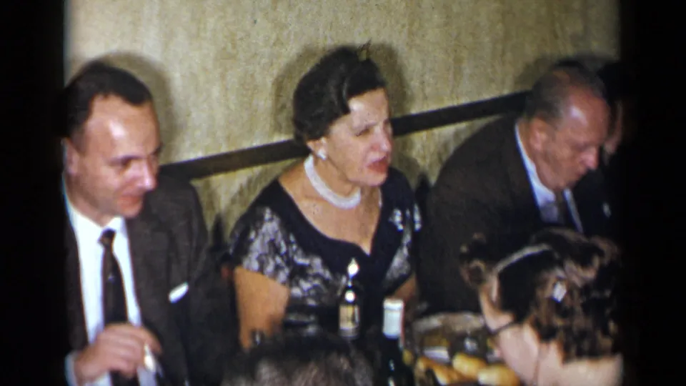1954 New York home-movie frame showing adults at a formal dinner table, including men in suits and a woman in a patterned black dress with pearls.