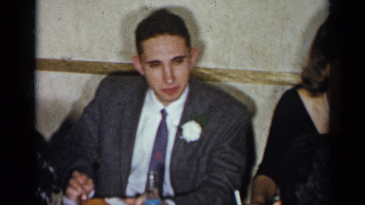 1954 New York home-movie frame with a young man in a gray suit and white boutonniere seated at a formal dinner table.