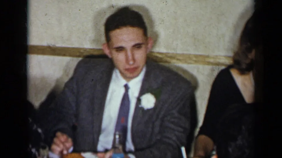 1954 New York home-movie frame with a young man in a gray suit and white boutonniere seated at a formal dinner table.