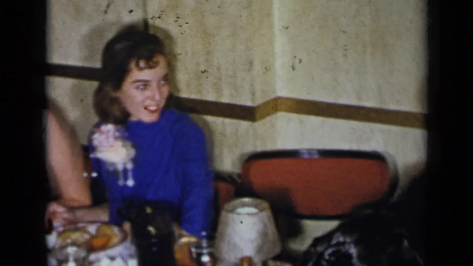 1954 New York home-movie frame showing a young woman in blue with a corsage seated at a dinner table in a banquet-room interior.